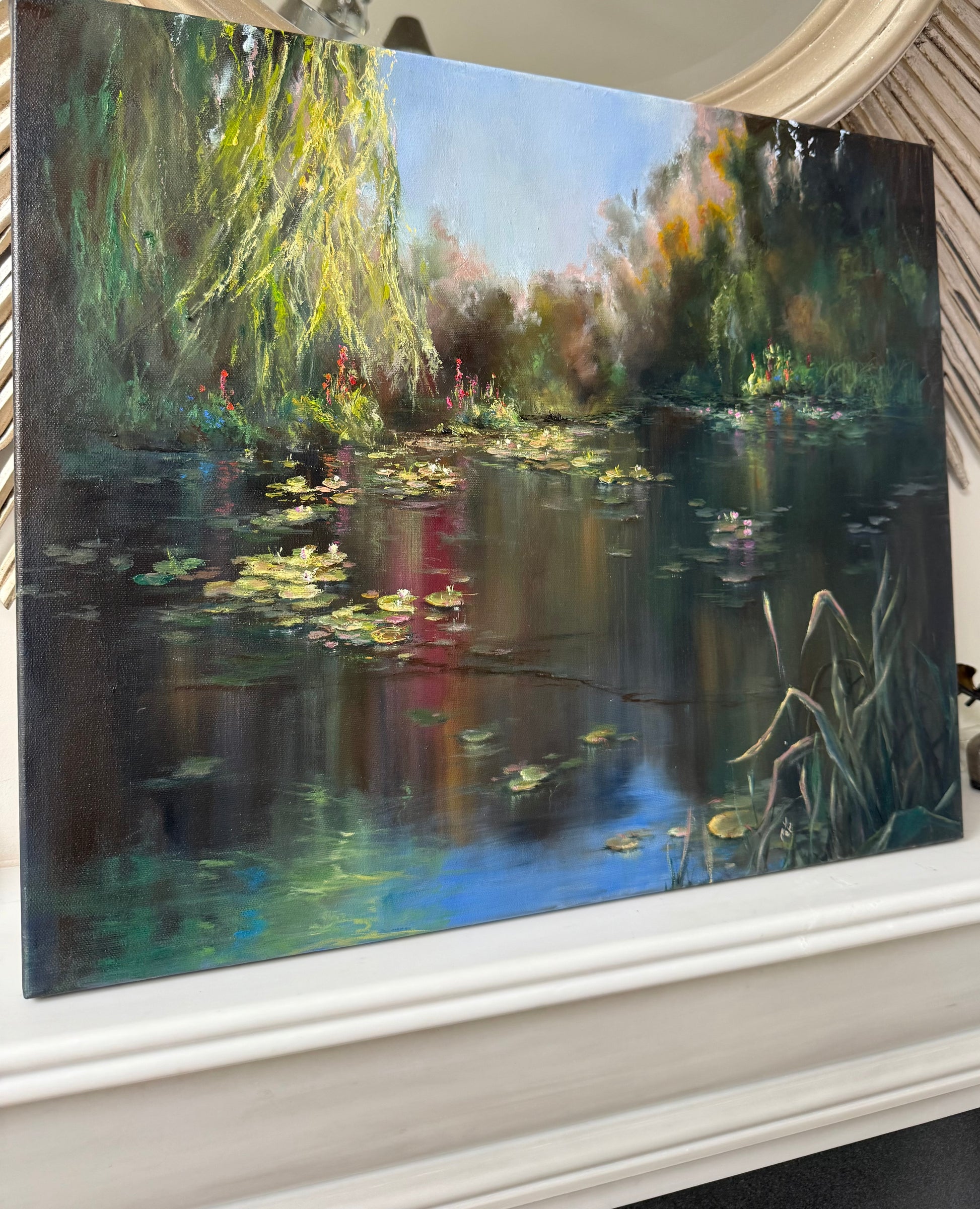 Lilies painting, showing a lily pond surrounded by willow trees. Sunlight is piercing through leaves highlighting lilies flowers and pads. Blue sky reflects on a dark pond water.