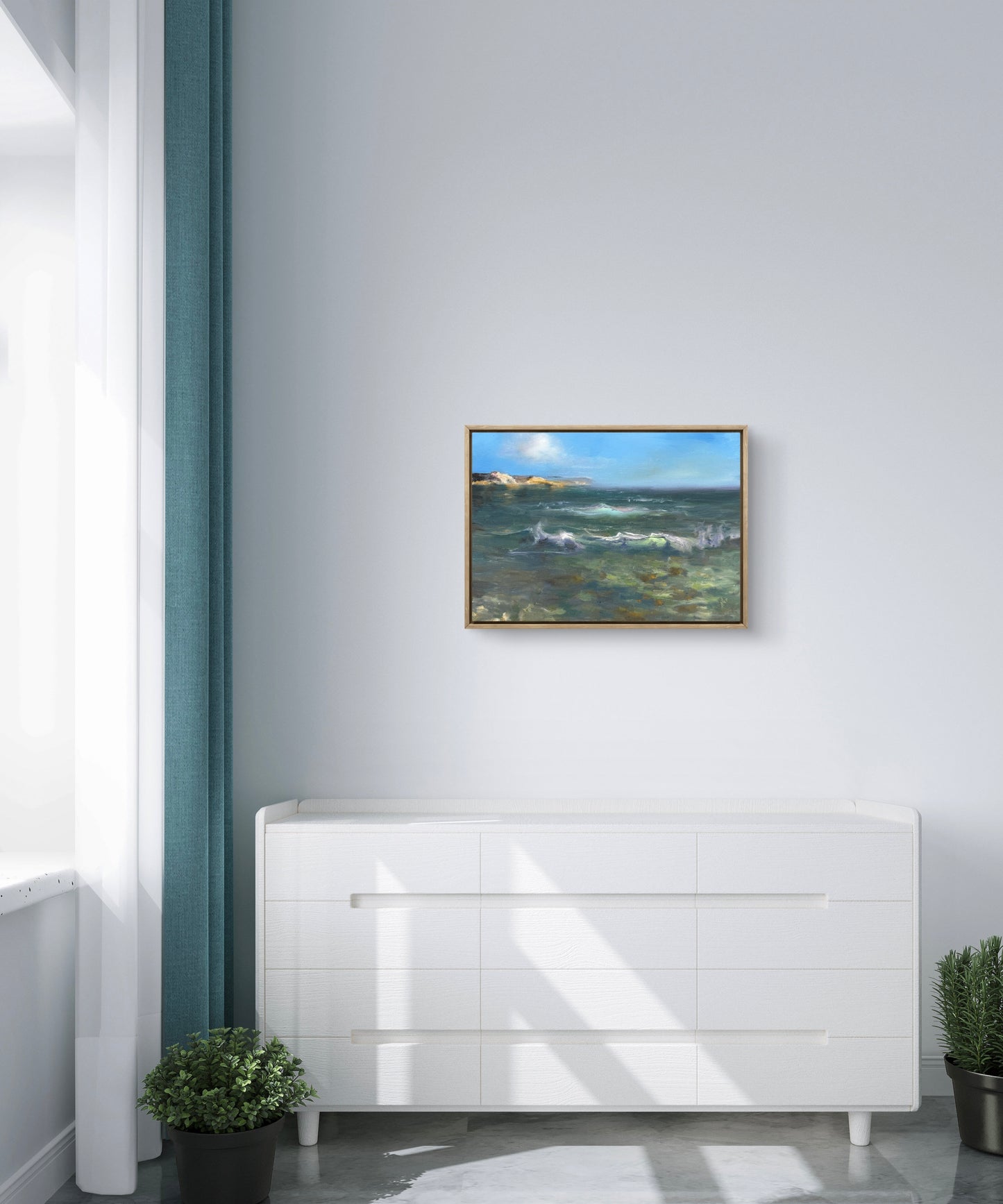 Framed ocean painting featuring blue sky, distant shores, turquoise waves and clear tidal waters in foreground. The seascape is hanging above a chest of drawers.