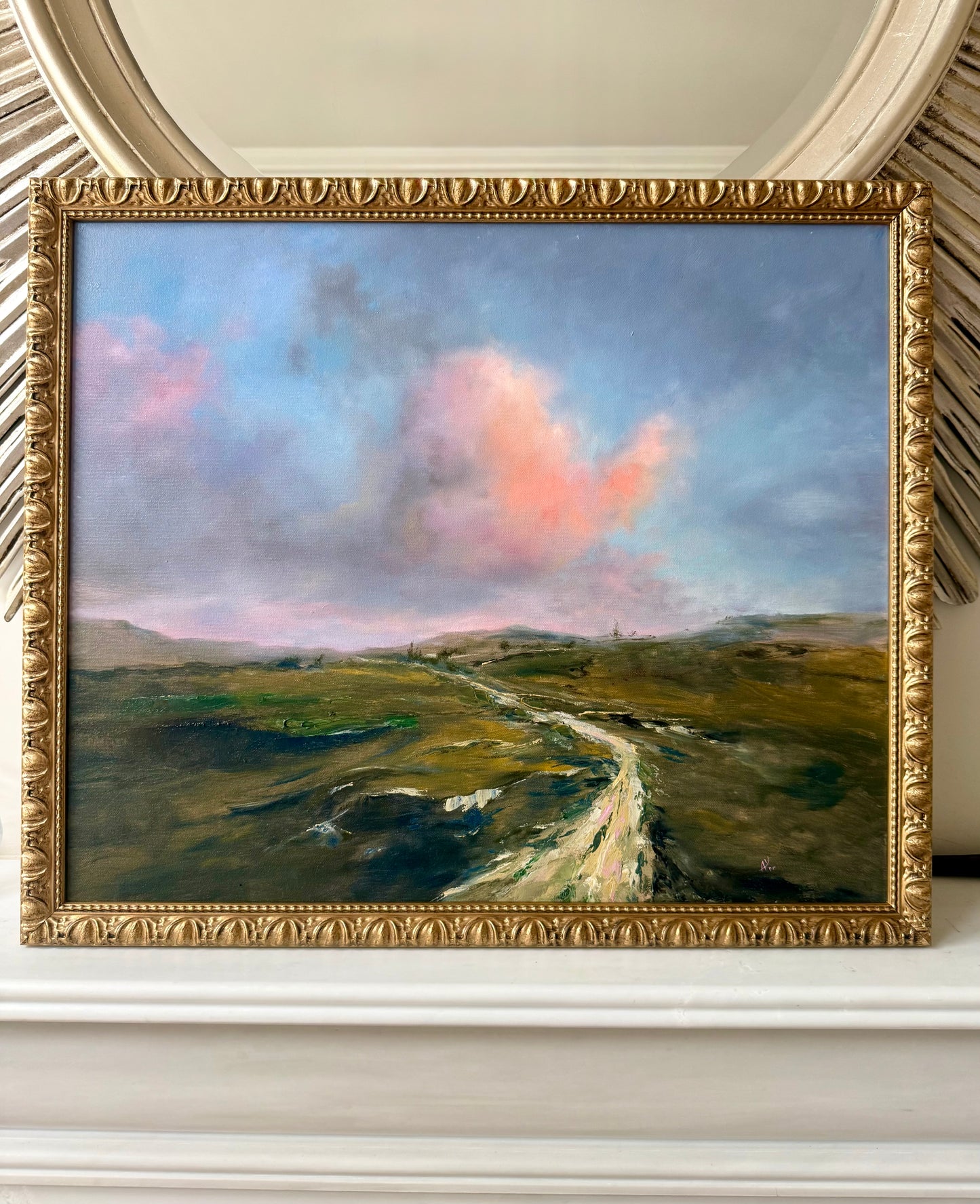 Framed landscape painting with pink clouds over green hills, with a gold frame on a white shelf above fireplace .
