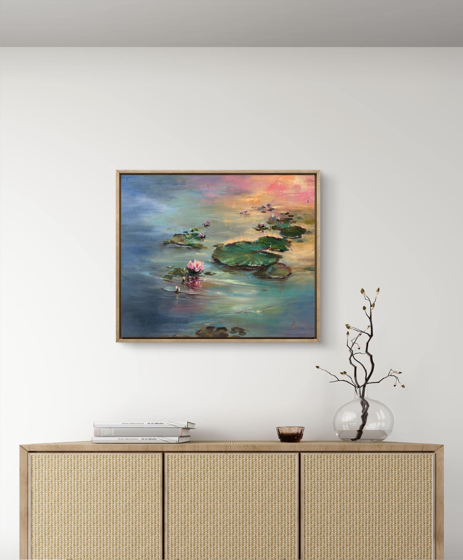 Framed painting of a serene landscape with water lilies on a wall above a wooden cabinet.