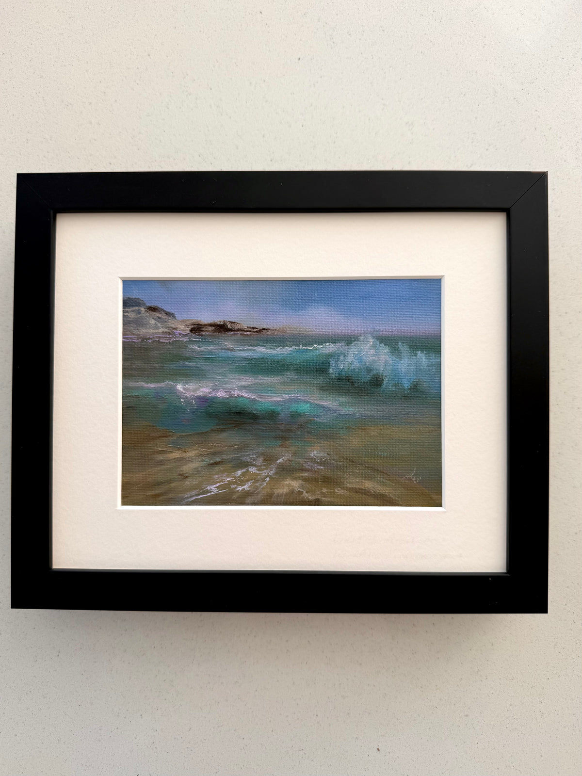 A framed coastal landscape painting featuring turquoise ocean waves and sandy shore, mounted with a wide white mat inside a black frame against a light textured wall.