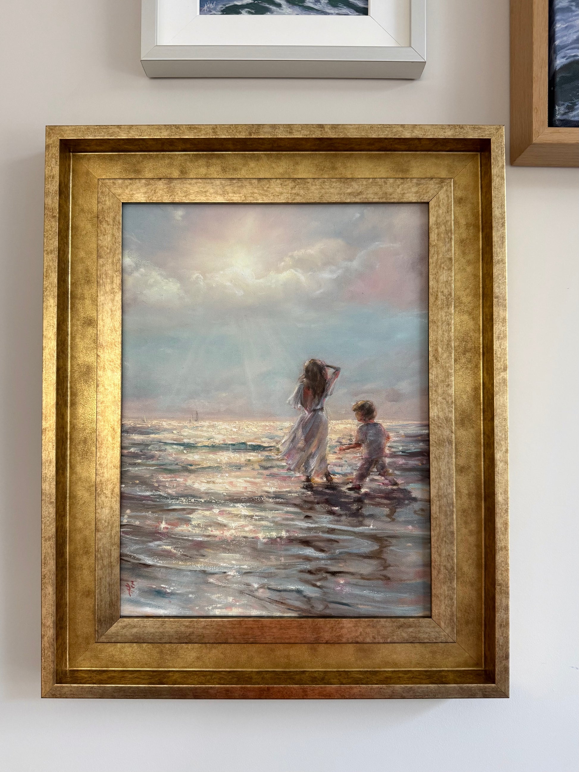 Framed painting of children playing on a beach with a gold frame hanging on the wall.