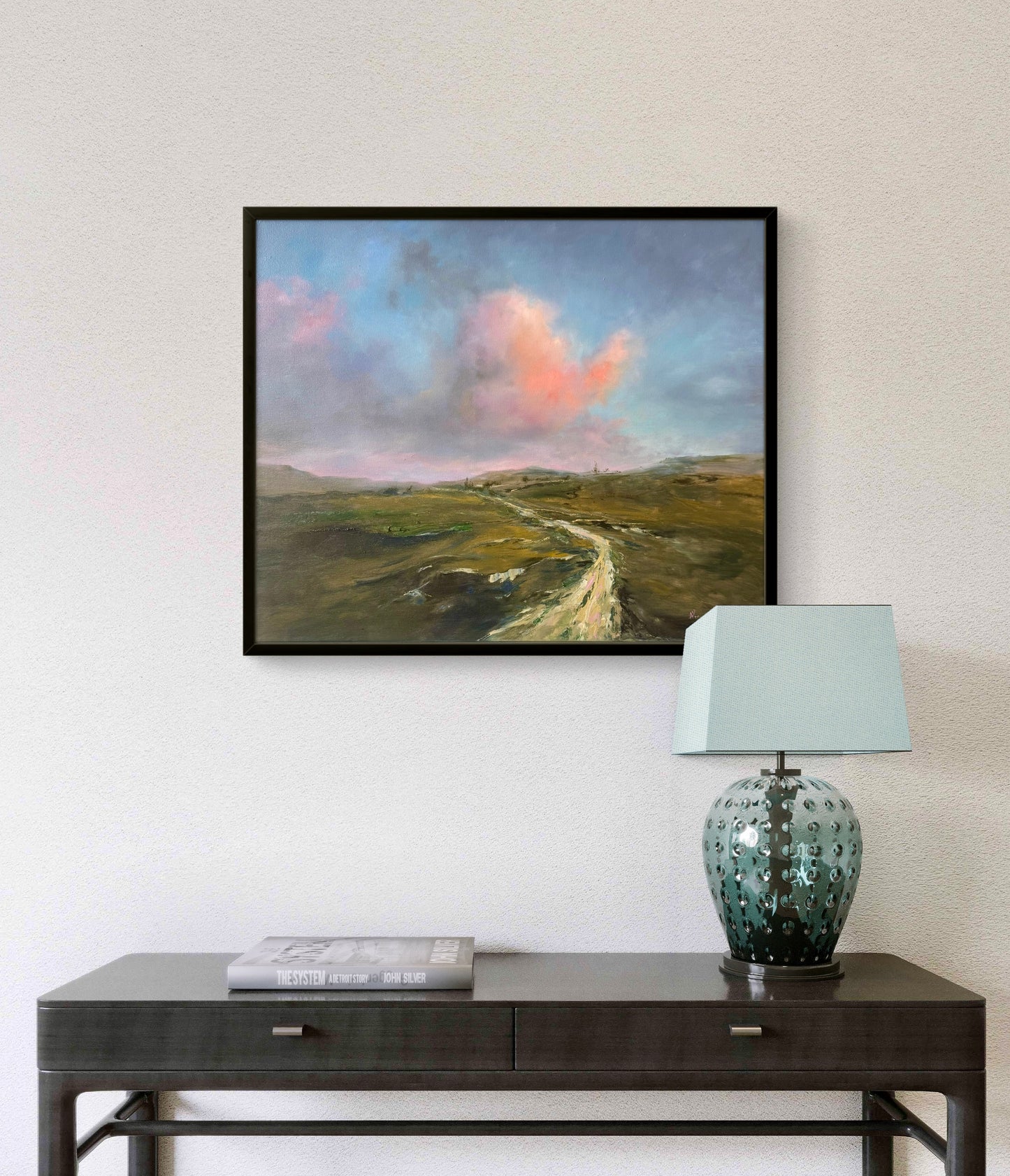 Framed pink and green  landscape painting above a dark wooden console table with a lamp.