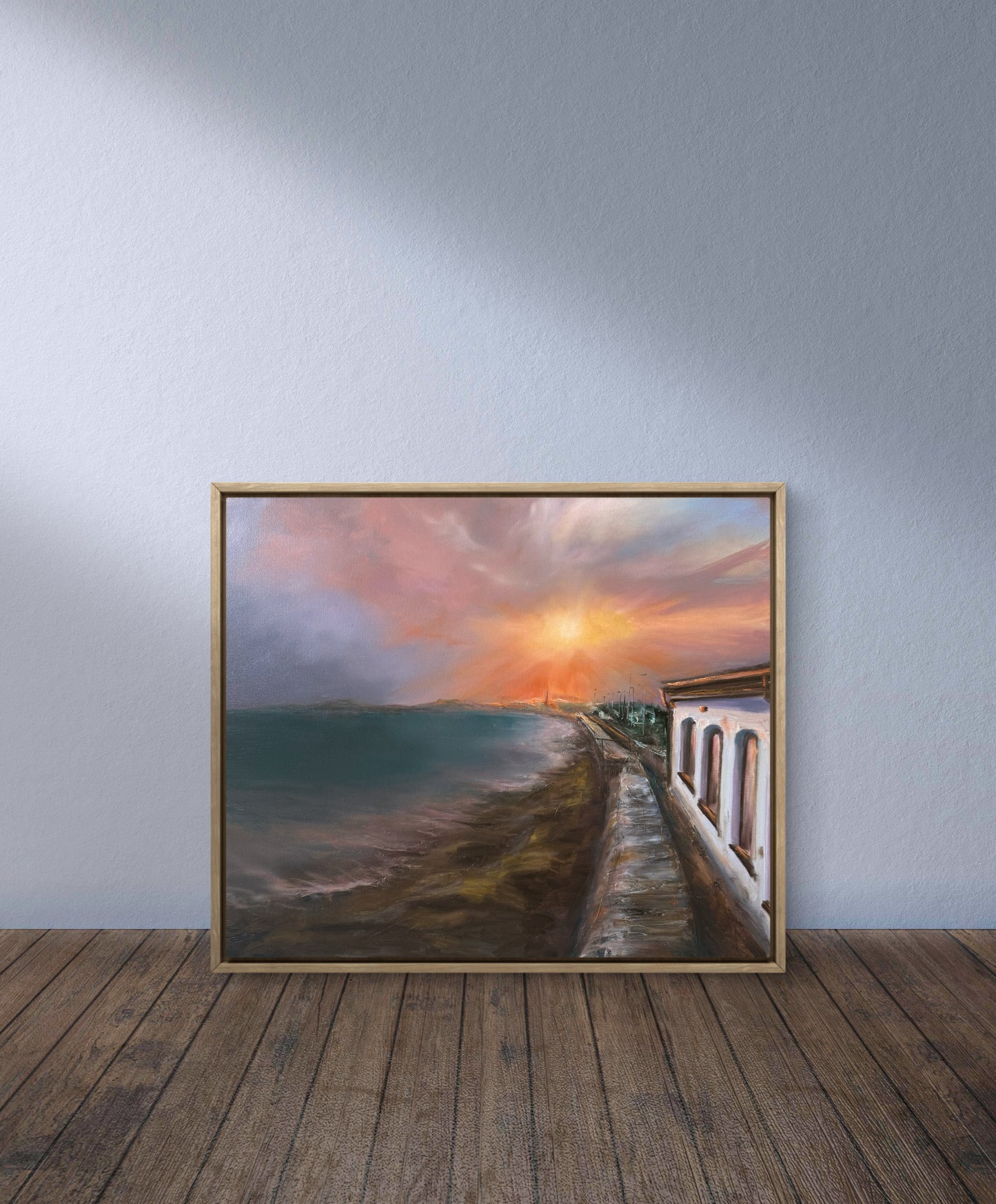 Framed artwork of a sunset over a coastal scene on a wooden floor with a light gray wall.