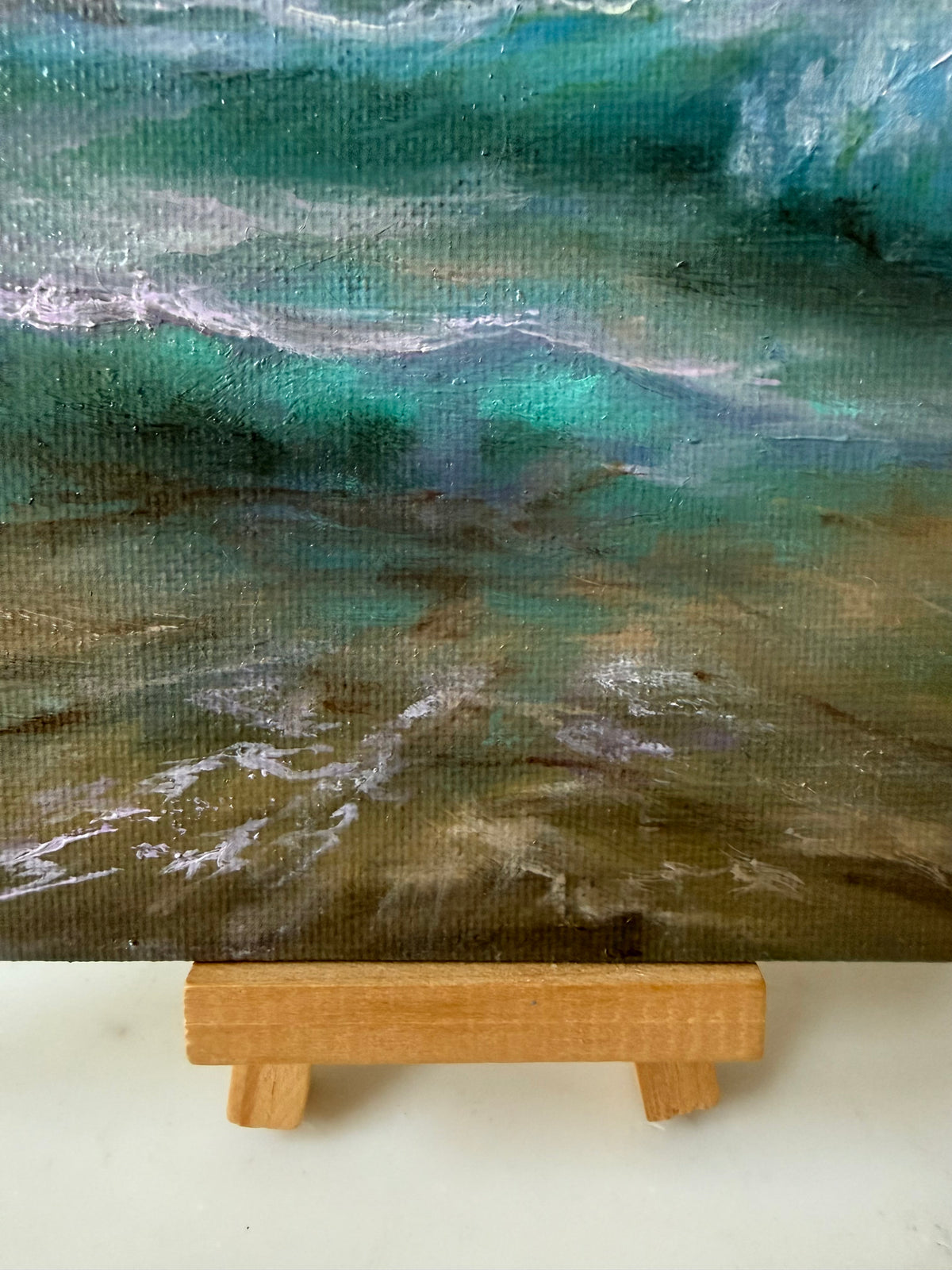 A close-up of a textured painting of ocean waves in shades of teal, green, brown, and white, displayed on a small wooden easel stand.