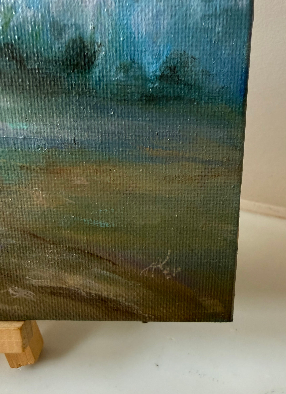 A close-up of a textured canvas painting showing a landscape scene with blue sky, green trees, and a brown field, displayed on a small wooden easel stand.