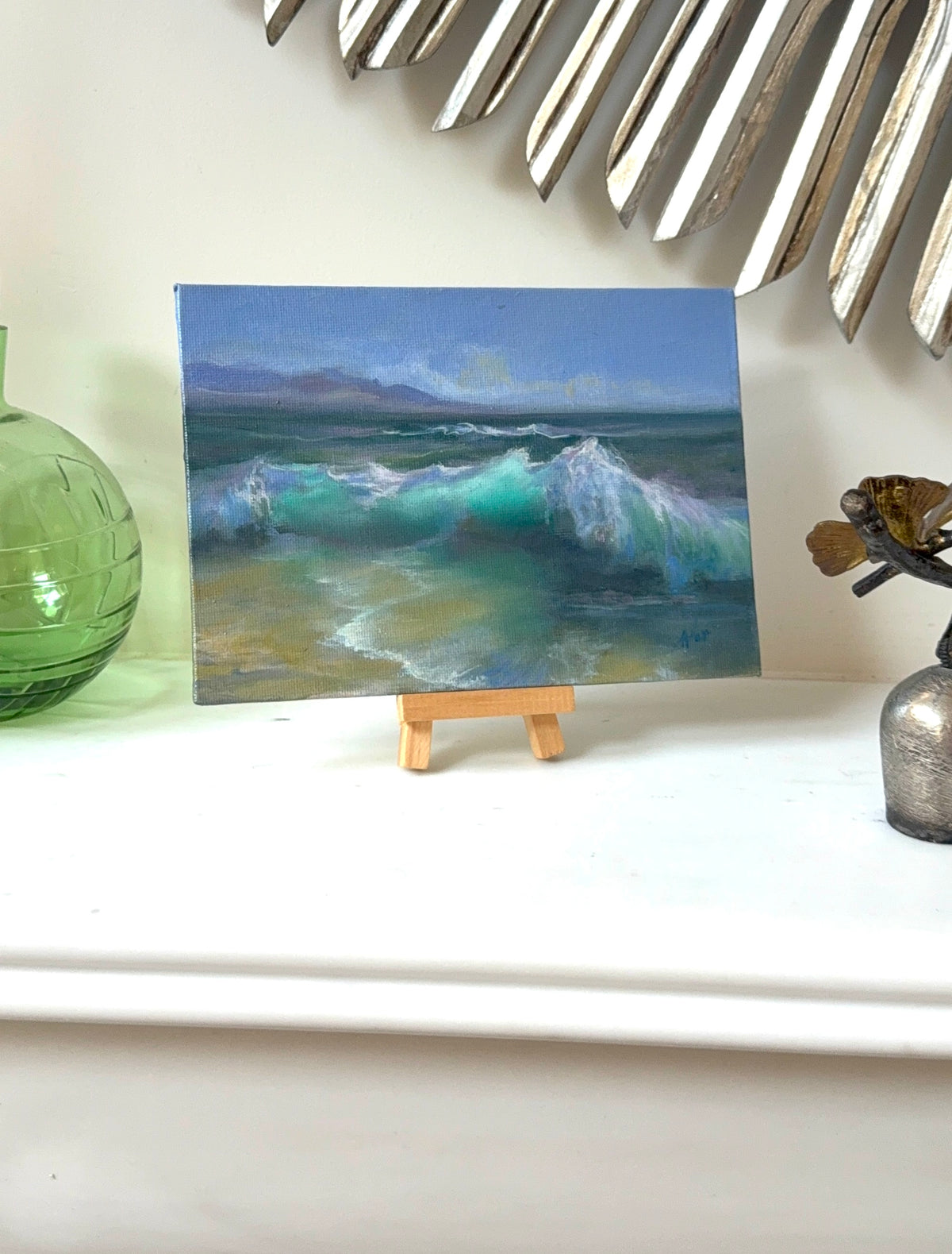 A small landscape painting of ocean waves displayed on a miniature wooden easel, set on a white shelf with decorative props including a green glass vase and a metallic wall mirror.