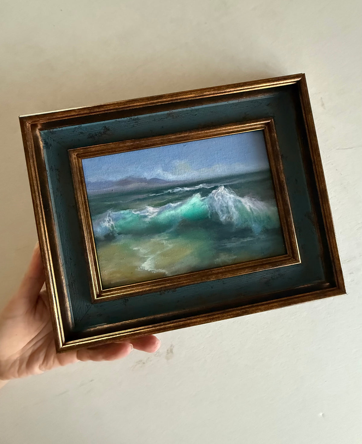 A hand holds a small framed painting of ocean waves with a distressed gold frame and dark teal mat against a plain off-white wall.
