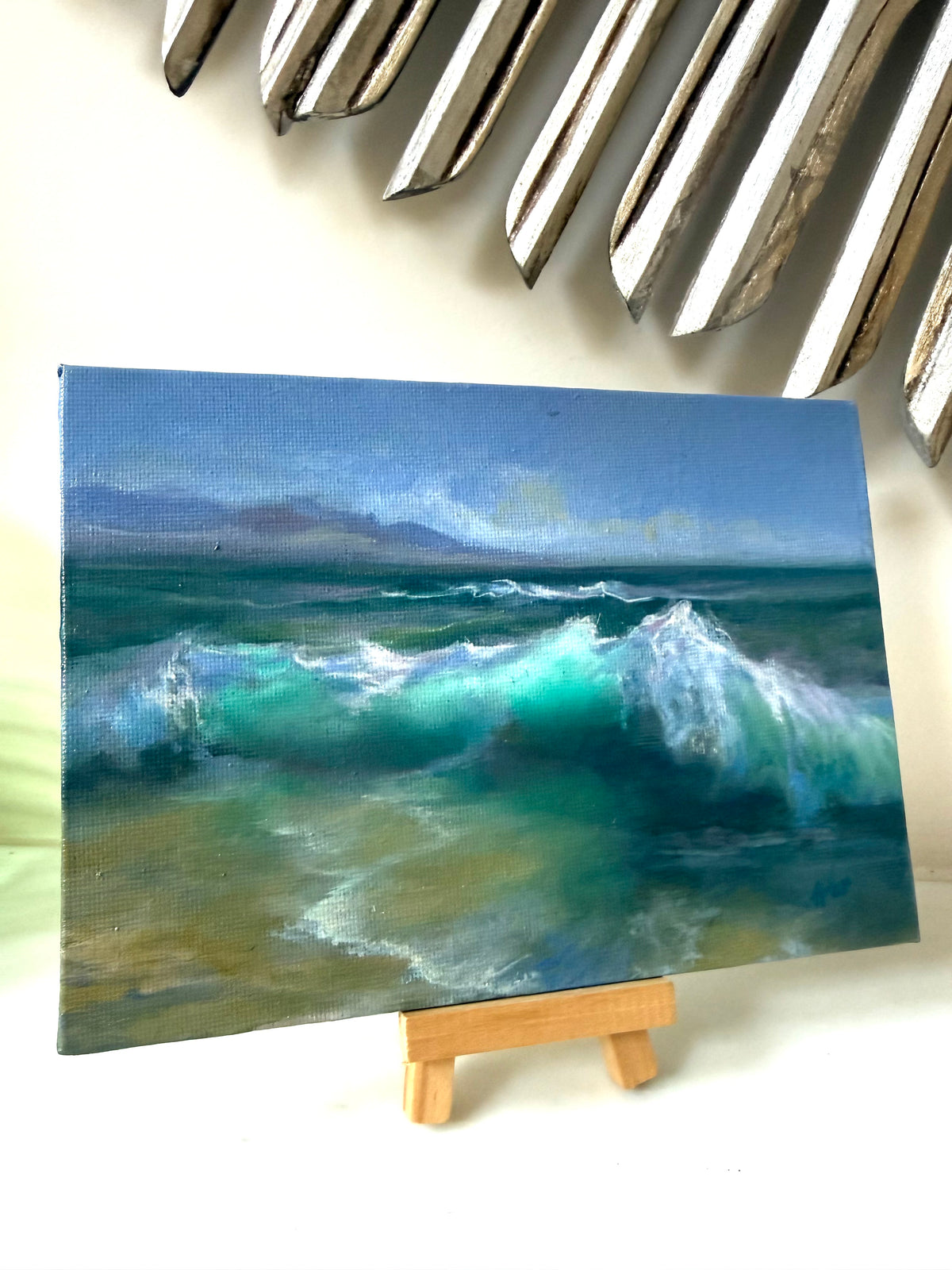 A small canvas painting of a turquoise and blue ocean wave, displayed on a miniature wooden easel against a light background with a decorative metallic wall piece above.
