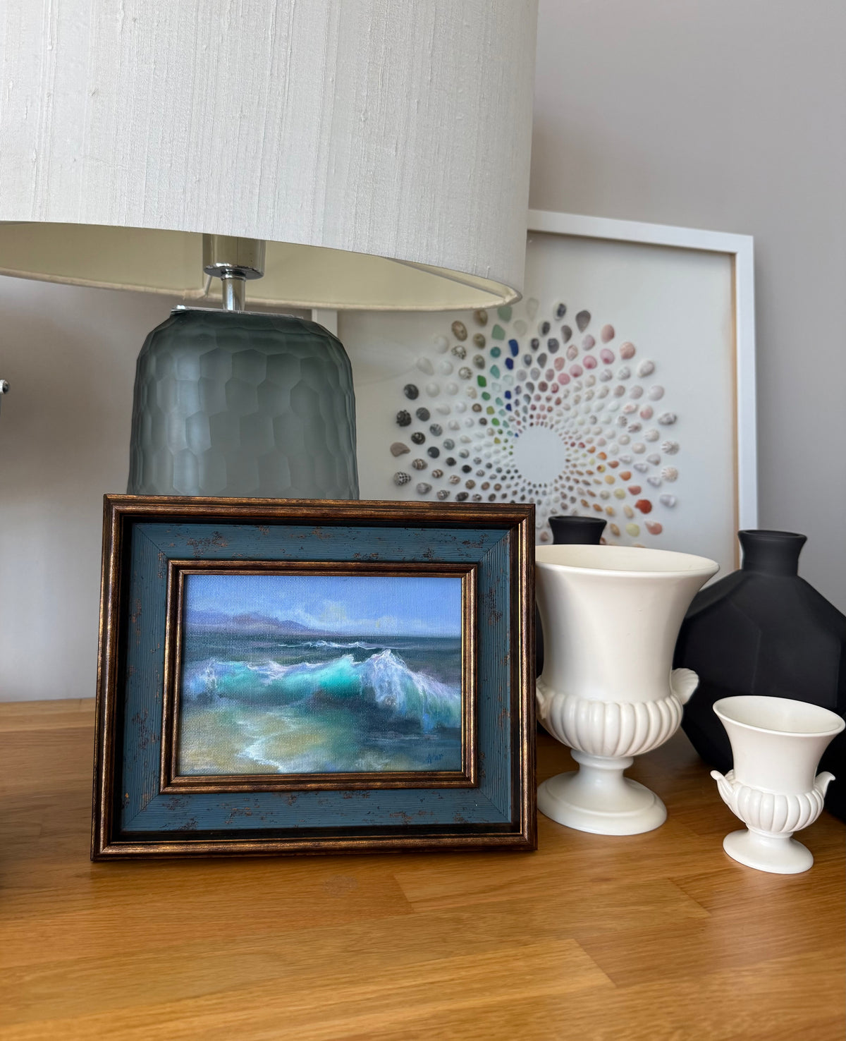 A framed square oil painting of a blue ocean wave scene, styled on a wooden table next to a lamp, white and black vases, and a large framed art print.