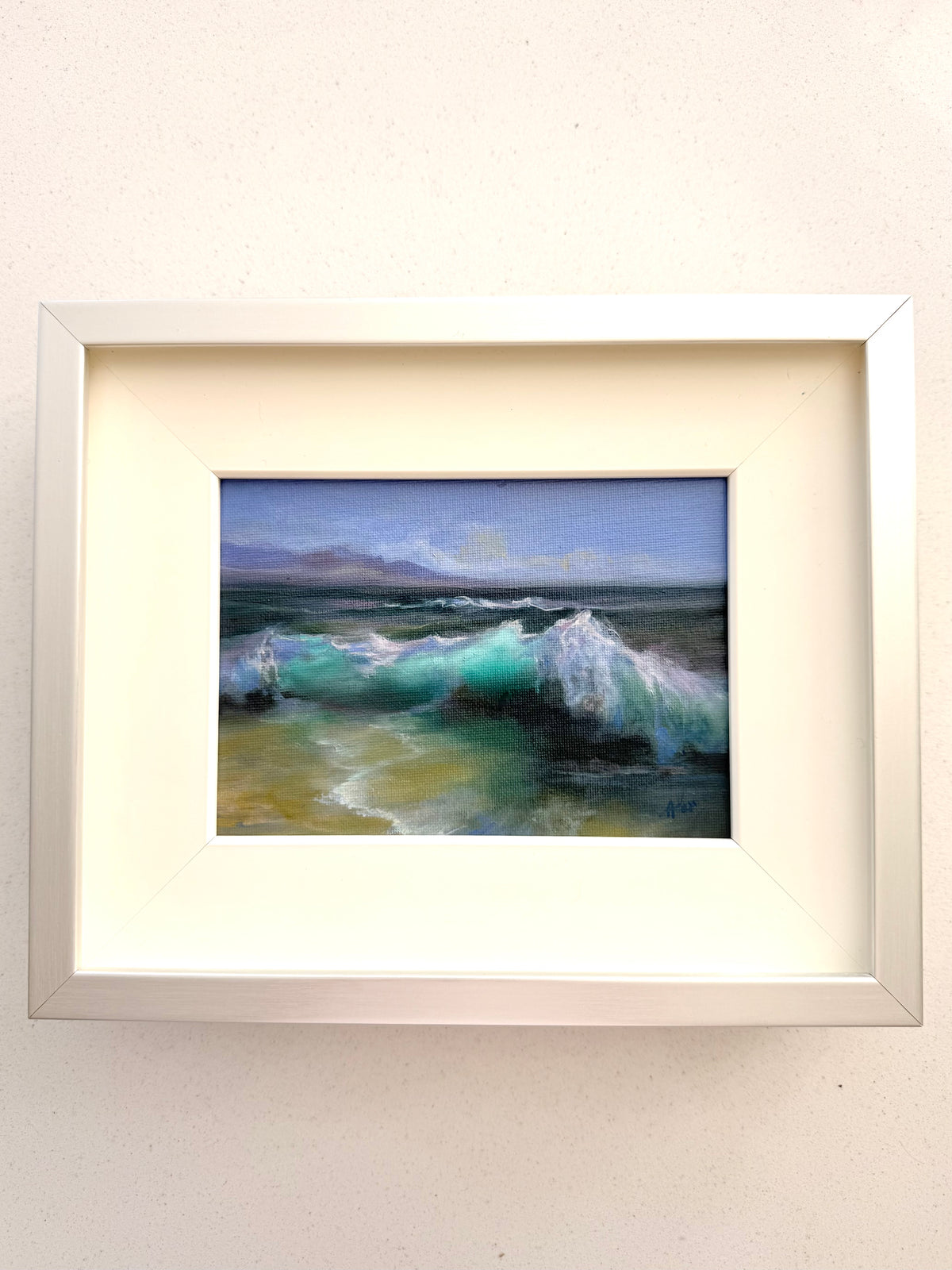 A framed landscape painting of ocean waves under a blue sky, presented in a white frame with a cream mat on a light beige wall.