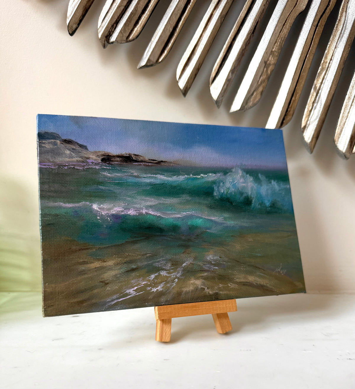 A small canvas painting of an ocean scene with turquoise waves and rocky shoreline, displayed on a miniature wooden easel against a light beige wall with decorative metallic leaves above.