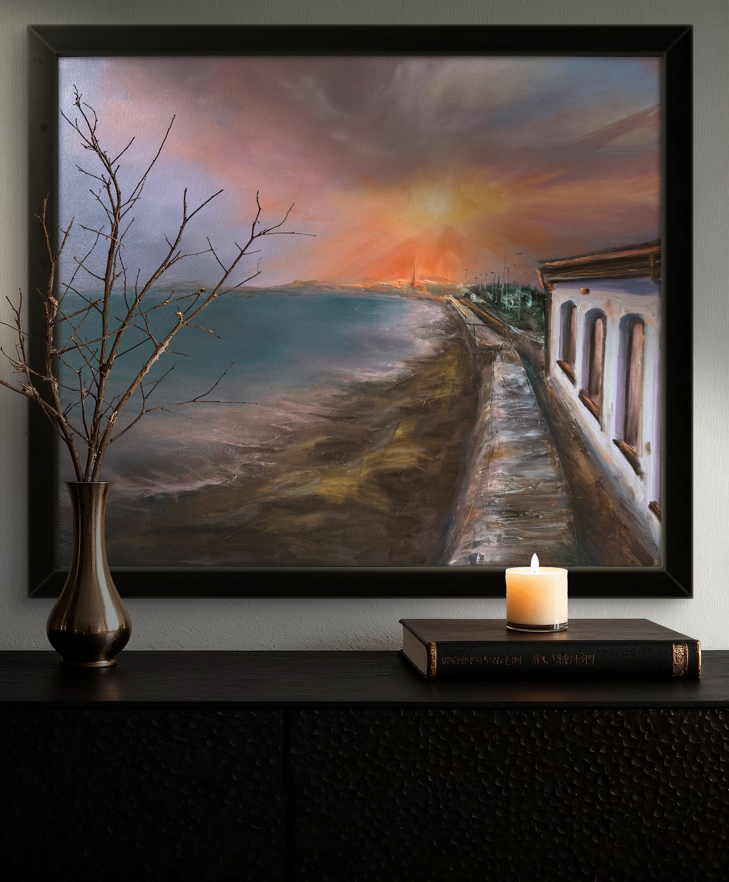Framed painting of a sunset over water with a candle and book on a shelf below.