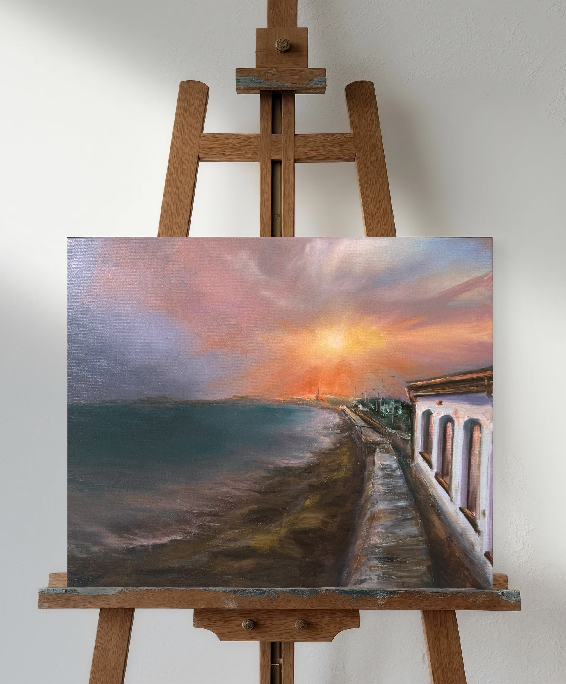 Painting of a sunset over a coastal scene on an easel against a white wall
