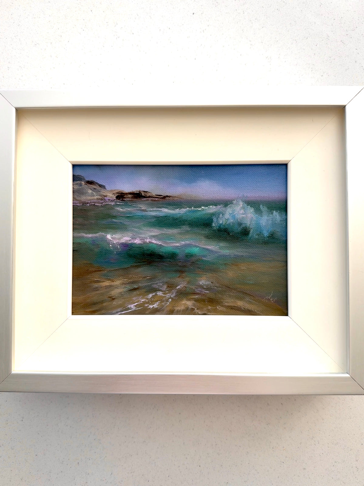 A framed landscape painting of a rocky beach and ocean waves, featuring a white frame and cream mat on a light textured wall.