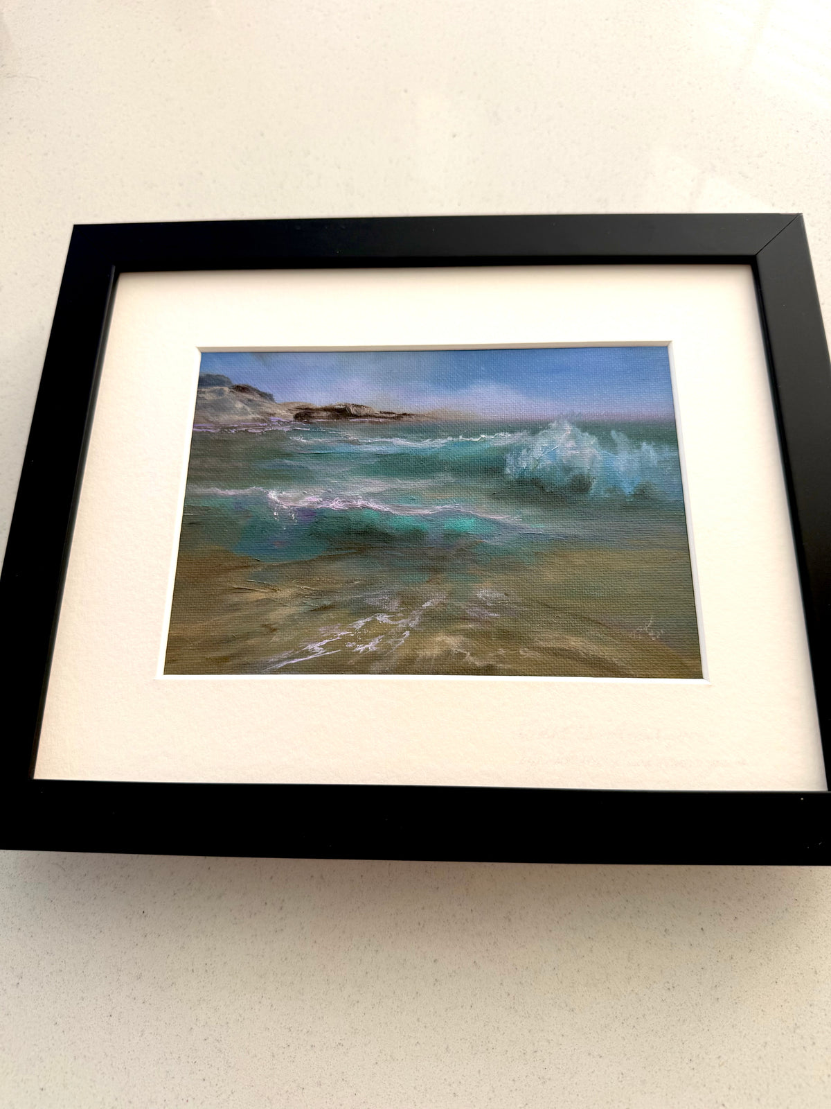 A framed coastal landscape painting featuring turquoise waves and rocky shoreline, presented with a wide white mat and a thick black frame on a light neutral background.