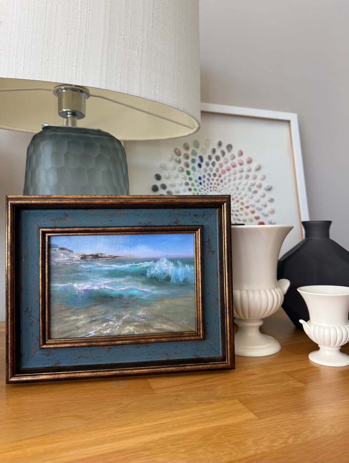 A framed coastal landscape painting with blue and white waves, displayed on a wooden table next to a blue-gray lamp base, white ceramic vases, and a dark vase.